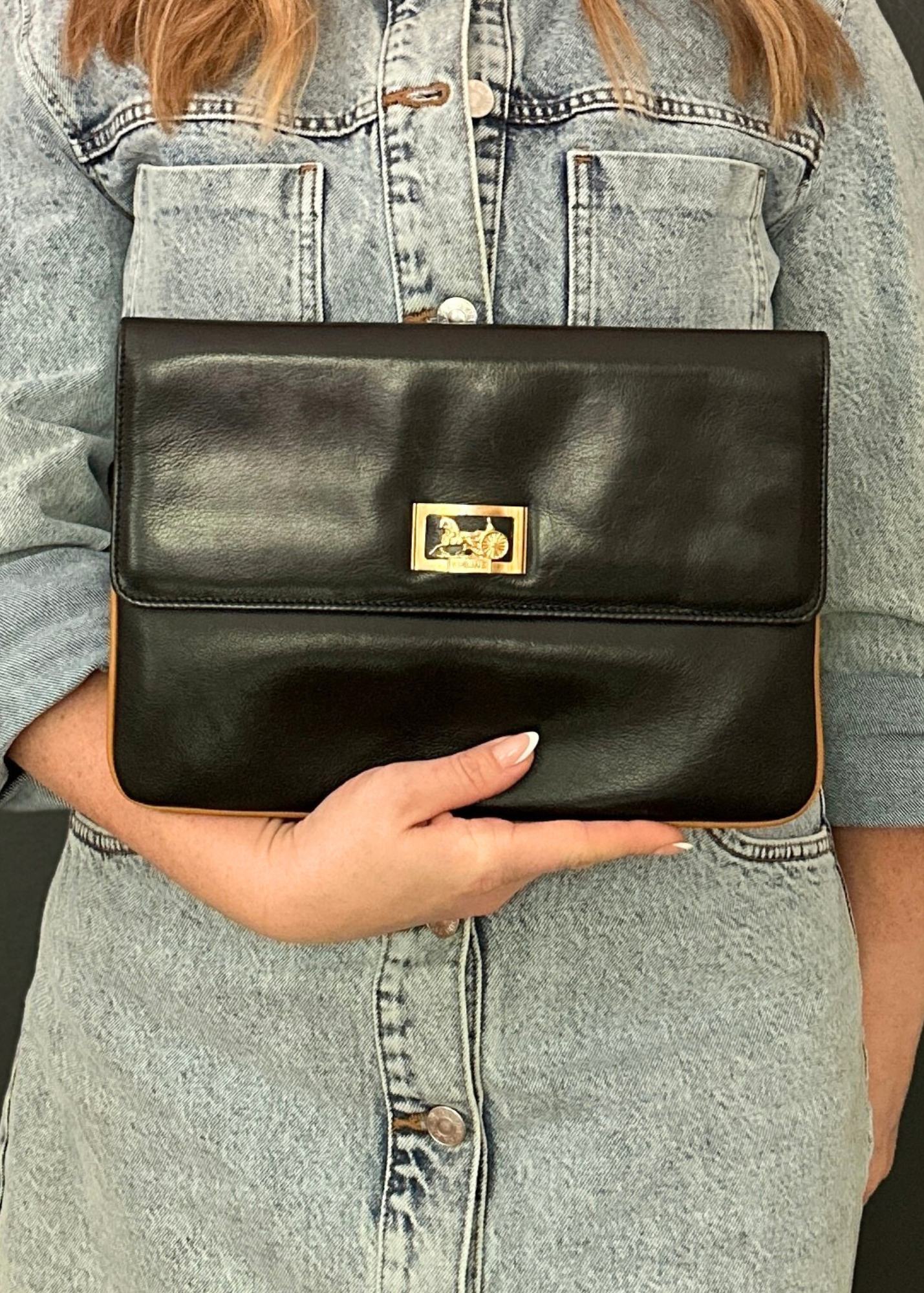 Person holding a black leather Celine clutch with a gold clasp against a denim background