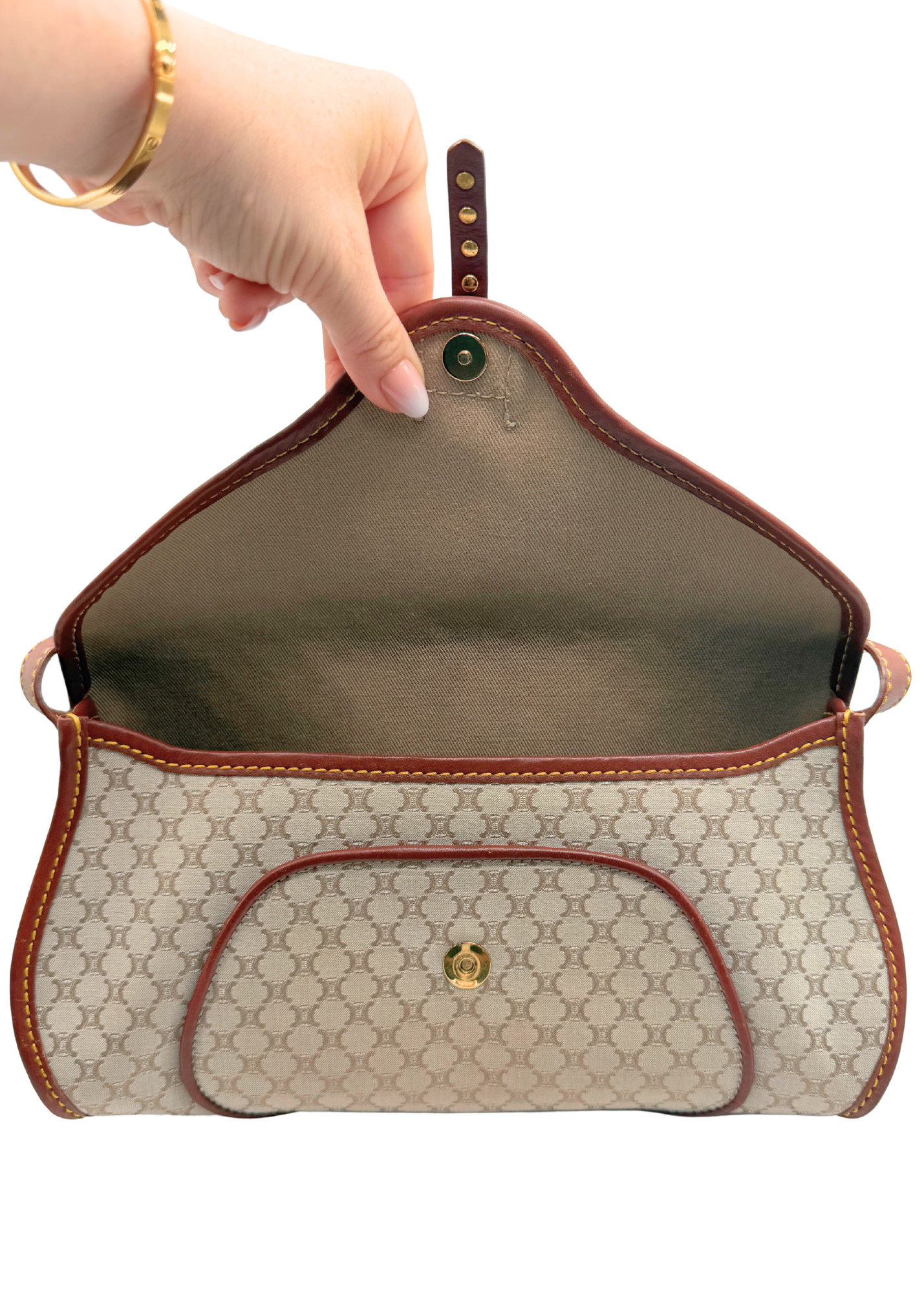 A beige and brown vintage Celine triomphe monogram handbag with a gold-tone logo clasp, held open by a hand wearing a gold bracelet against a white background.
