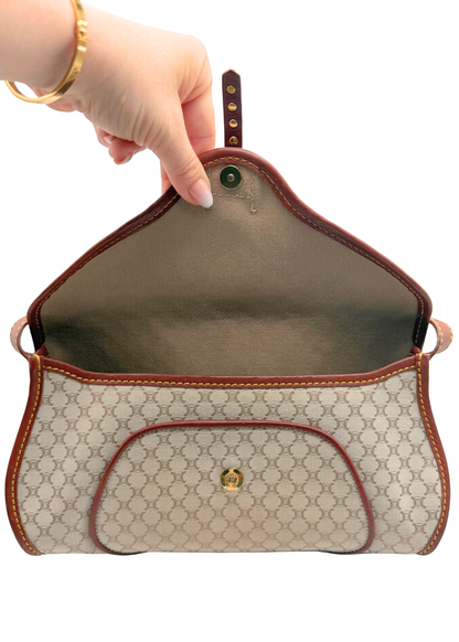A beige and brown vintage Celine triomphe monogram handbag with a gold-tone logo clasp, held open by a hand wearing a gold bracelet against a white background.