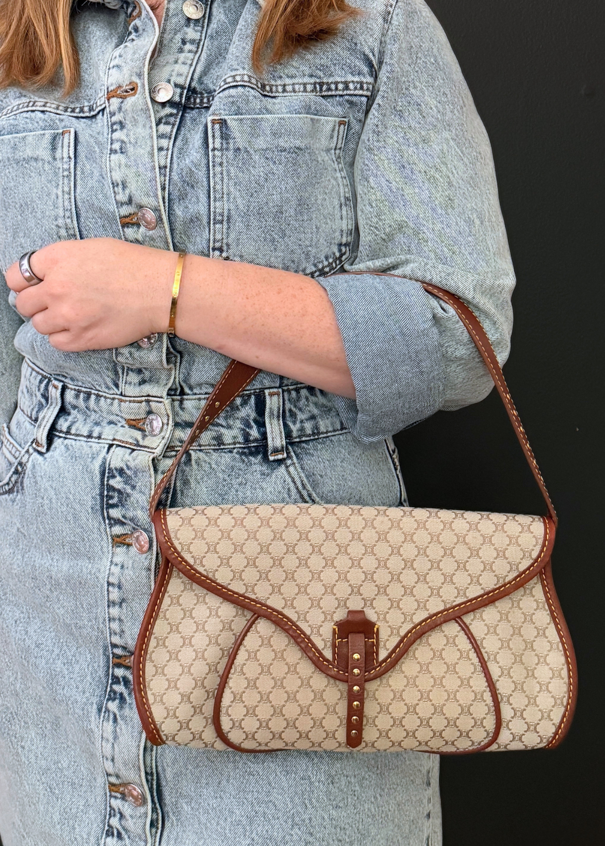 A person wearing a light blue denim jacket holds a vintage Celine beige and brown patterned shoulder bag with a flap closure and brown leather trim against a dark background.