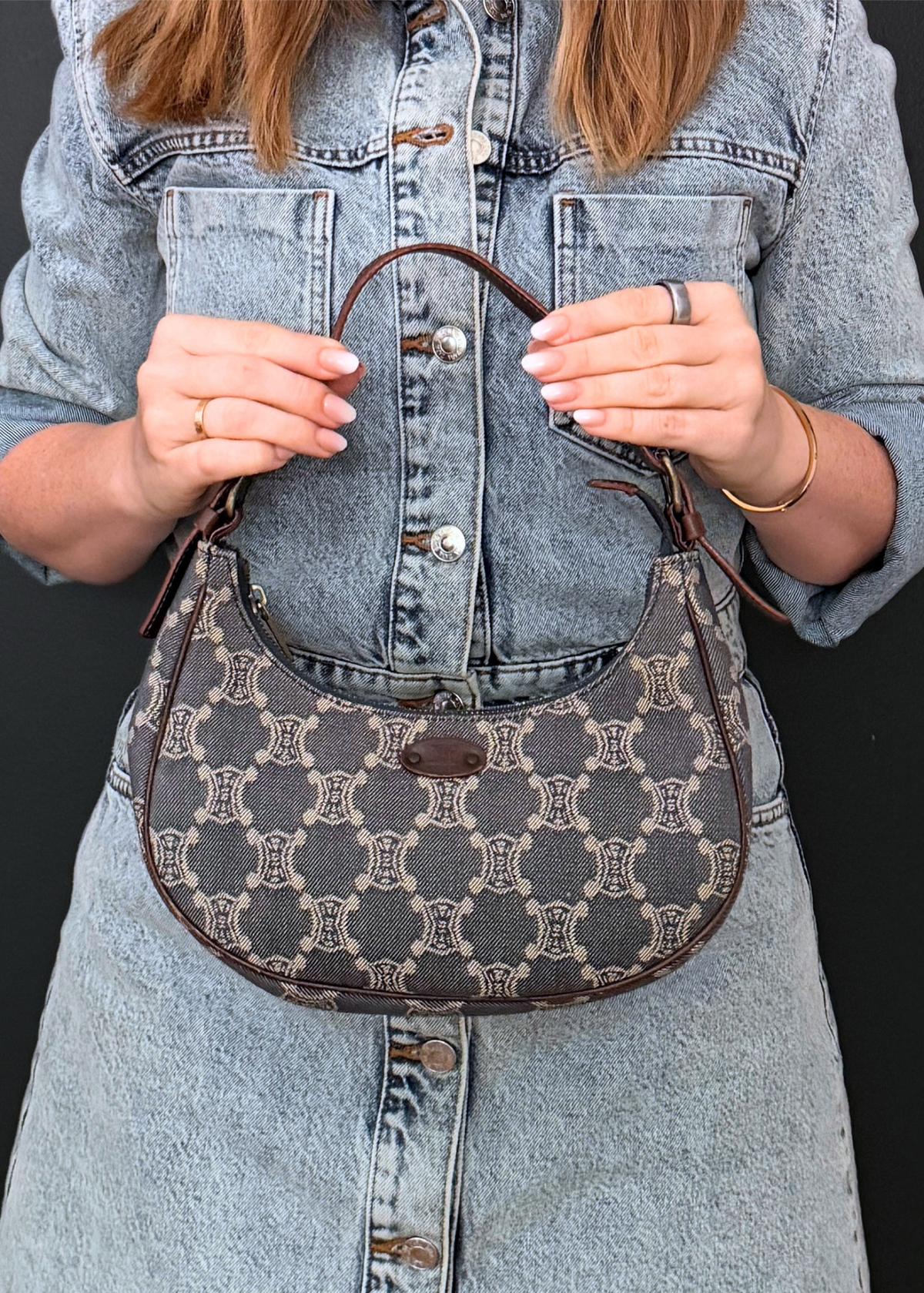 A person wearing a denim shirt holds a small vintage Celine crescent-shaped Ava shoulder bag with a brown monogram pattern and dark brown leather trim against a dark background.