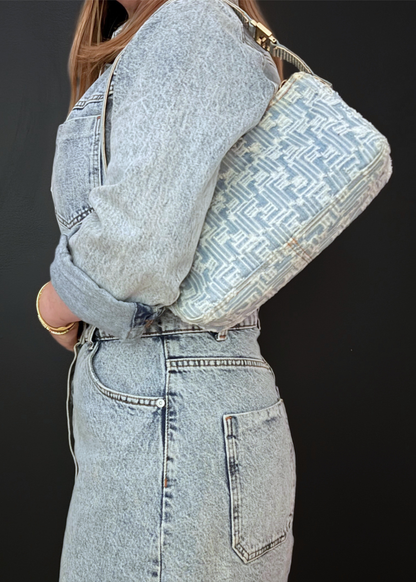 A person wearing a light-wash denim jacket and matching denim skirt carries a vintage Fendi denim shoulder bag with a blue and white Zucca pattern and gold-tone hardware against a dark background.