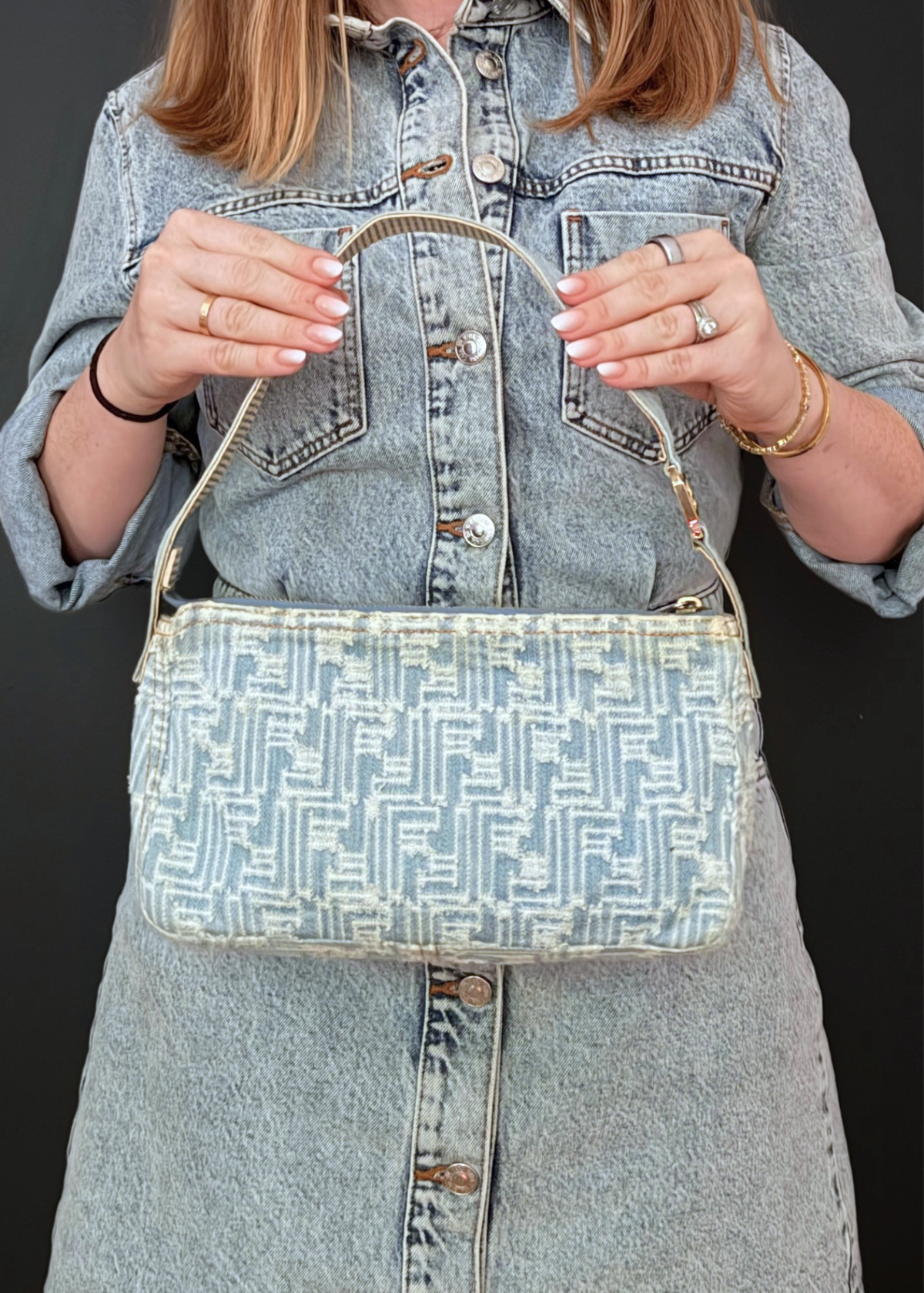 A person wearing a light-wash denim shirt holds a vintage Fendi denim handbag with a blue and white geometric monogram pattern and a gold-tone top handle against a dark background.