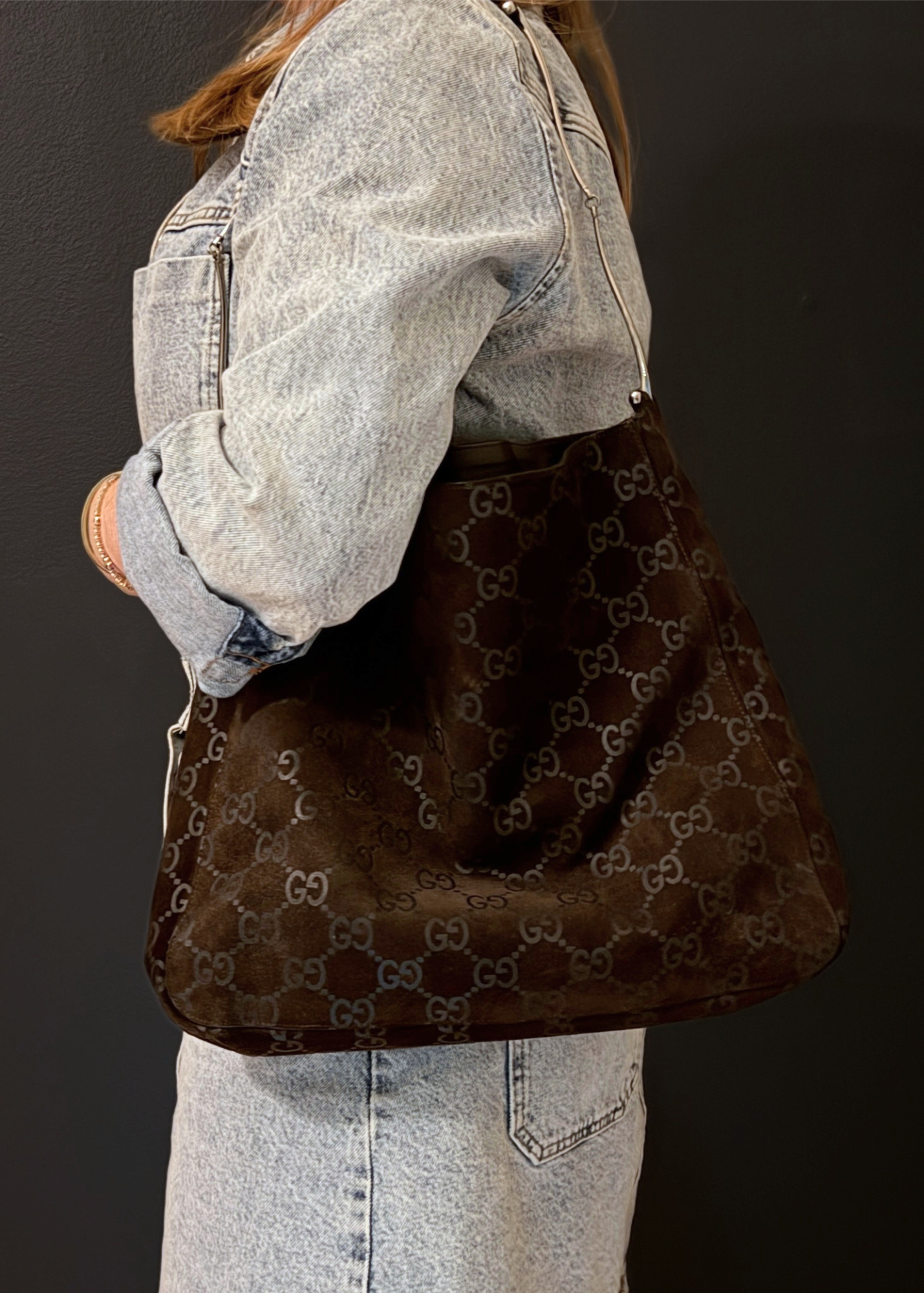 A person wearing a light-wash denim jacket and skirt carries a large brown monogram Gucci shoulder bag with a dark brown suede-like front panel, photographed against a dark studio background.