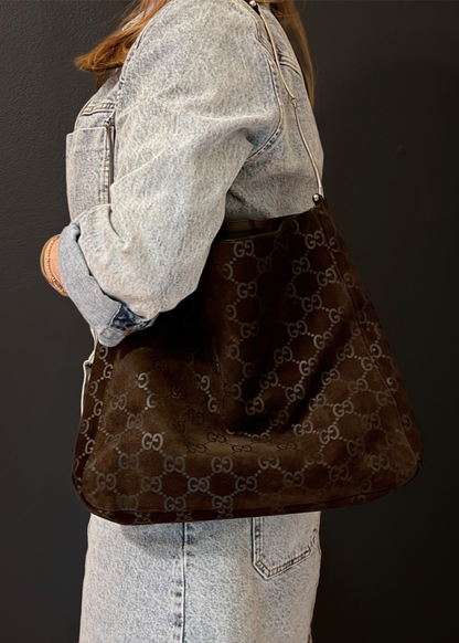 A person wearing a light-wash denim jacket and skirt carries a large brown monogram Gucci shoulder bag with a dark brown suede-like front panel, photographed against a dark studio background.