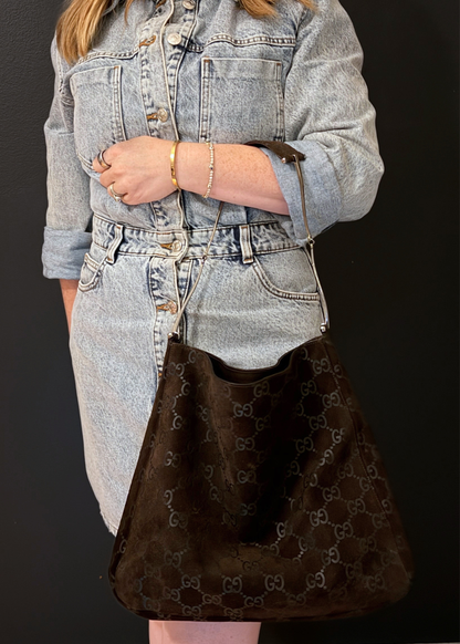 A person wearing a light-wash denim dress or outfit holds a large dark brown Gucci tote bag with the brand's repeating GG monogram pattern against a dark background.