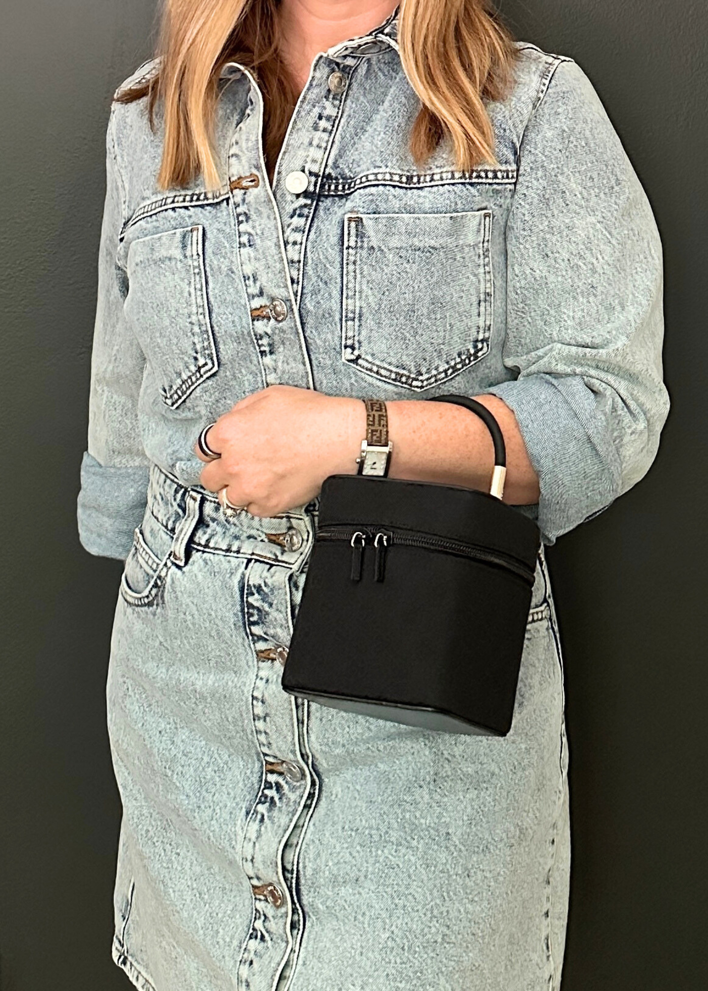 Person wearing a light blue denim dress holding a black handbag against a dark background