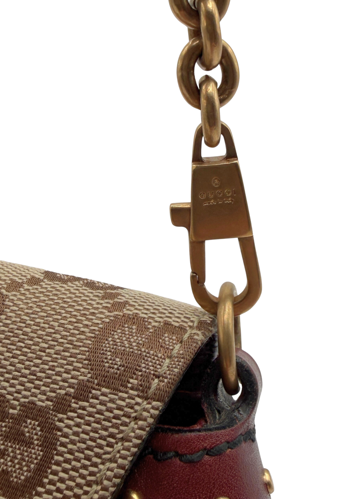 Close-up of a Gucci handbag corner showing the brown and beige monogram canvas, dark red leather trim, and gold-tone hardware clasp on a white background.