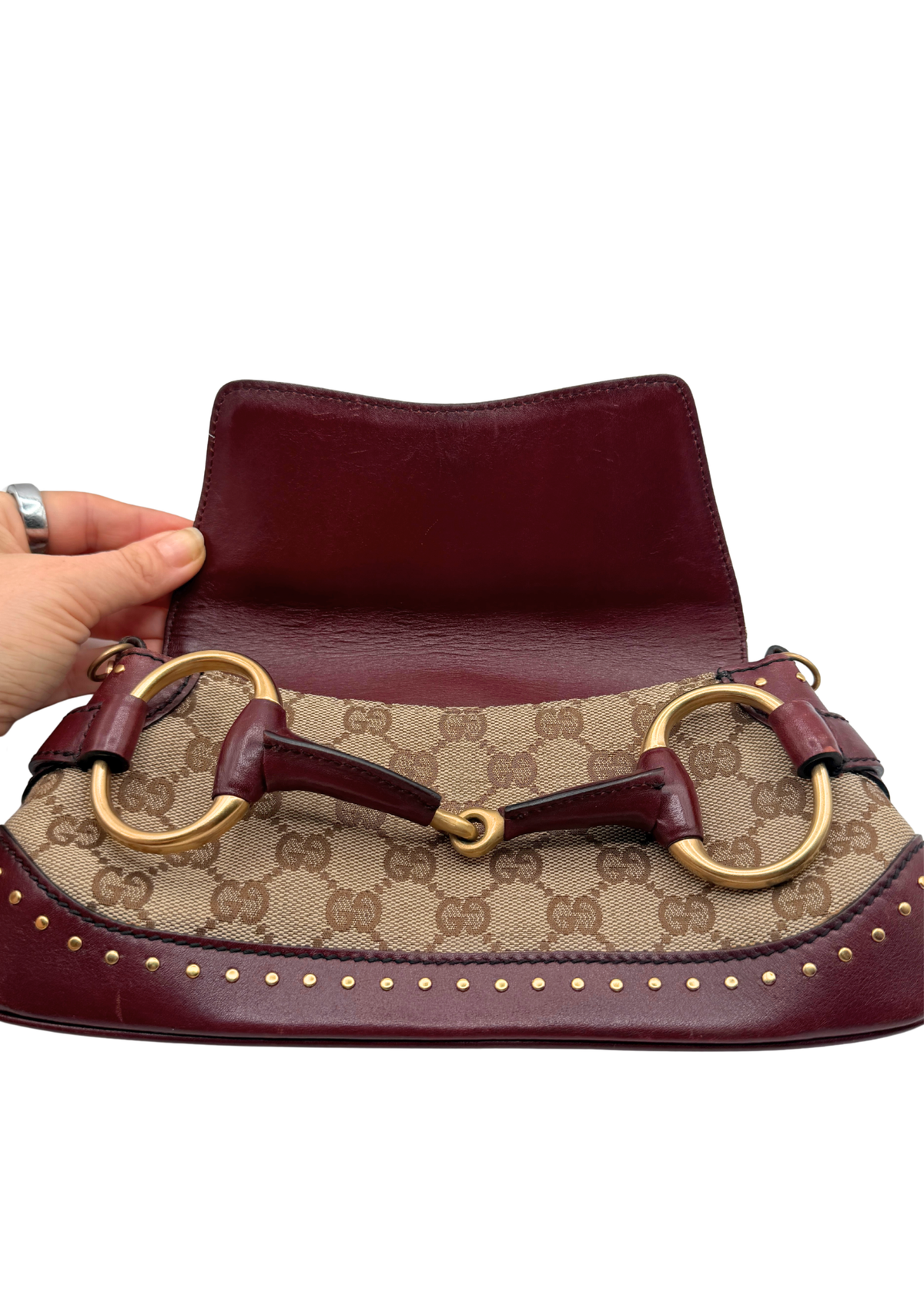 A Gucci Horsebit shoulder bag in beige GG monogram canvas with burgundy leather trim and gold-tone hardware, held open by a hand against a white background.