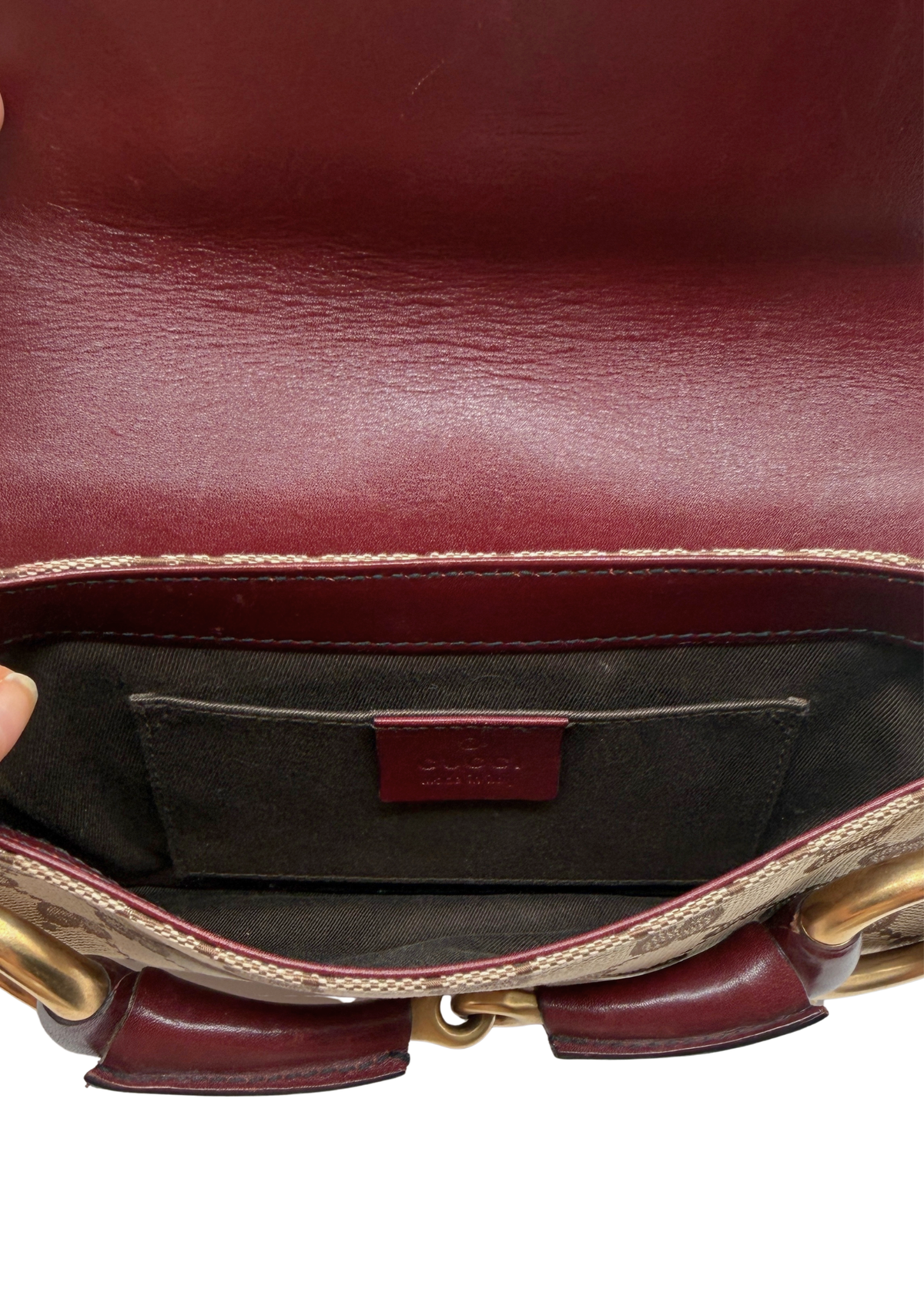 Close-up of the interior of a Gucci shoulder bag, showing a burgundy leather interior lining, a dark brown patterned fabric pocket with a small burgundy Gucci label, and gold-tone chain details at the bottom.