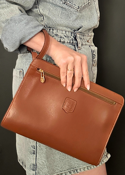 Brown leather clutch held by a person wearing a gray jacket and denim jeans.