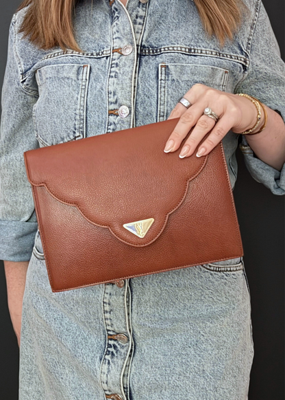 A person in a light blue denim shirt holding a vintage brown pebbled leather clutch YSL bag with a scalloped flap and a small gold-tone triangular logo plaque.