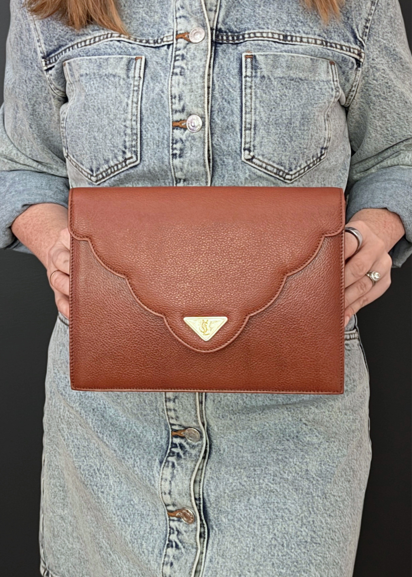 A person wearing a light-wash denim shirt holds a brown vintage textured leather YSL clutch bag with a scalloped flap and a small gold-tone triangular logo plaque.