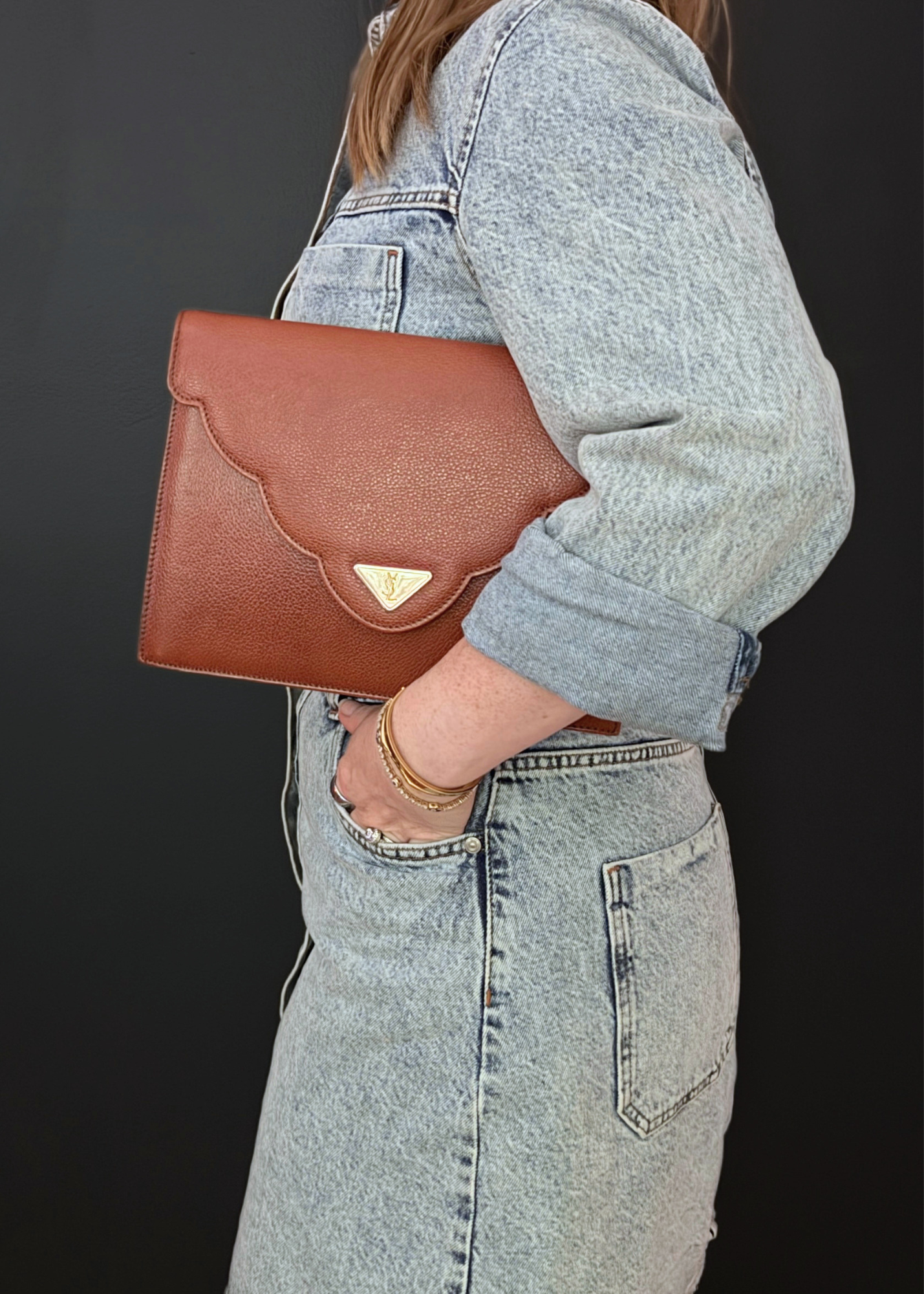A person wearing a light-wash denim jacket and matching denim skirt carries a vintage brown textured leather YSL clutch bag with a scalloped flap and a small gold-tone triangular logo plaque, set against a dark background.