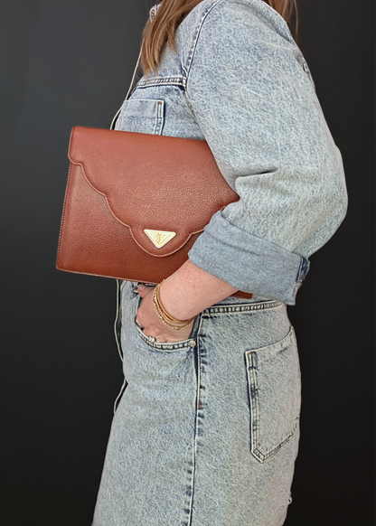 A person wearing a light-wash denim jacket and matching denim skirt carries a vintage brown textured leather YSL clutch bag with a scalloped flap and a small gold-tone triangular logo plaque, set against a dark background.