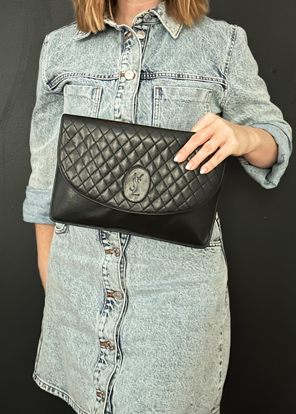 Person holding a black quilted YSL clutch bag against a dark background